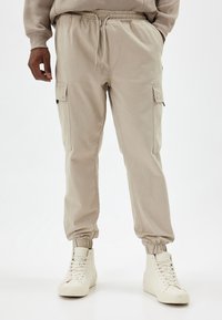 Beiga cargobyxor med elastiska avslut och midja, utrustade med sidofickor och ett smidigt, lätt tyg. Kombinationen med vita högknäliga sneakers.