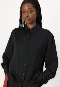 Camisa preta de botão com gola estruturada, mangas longas e uma textura lisa e elegante. Apresenta botões pretos e detalhes de costura discretos.
