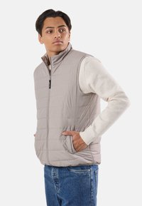 Młody mężczyzna w jasnobeżowej pikowanej kamizelce na kremowym swetrze, w niebieskich dżinsach, z jedną ręką w kieszeni kamizelki, patrzący na bok.