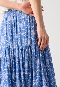 Blaue, gestufte Maxiröcke mit weißen Blumenmuster, ausgestattet mit einem gerüschten Bund und aus leichtem Baumwollmaterial. Sichtbare Handplatzierung an der Taille.