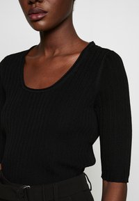 Mustapohjaista ribbikudonta yläosaa, jossa on pyöreä kaula-aukko ja puolihihat. Yläosassa on teksturoitu pinta ja tiukka siluetti.