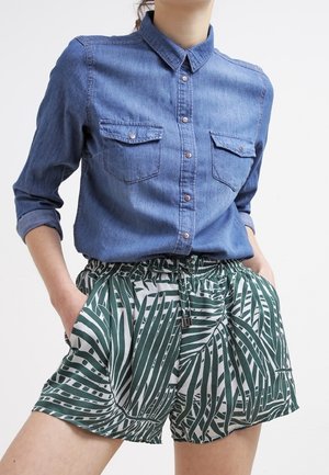 Persona que lleva una camisa vaquera azul abotonada con las mangas remangadas y pantalones cortos con patrón verde y blanco y cintura con cordón, con las manos en los bolsillos.