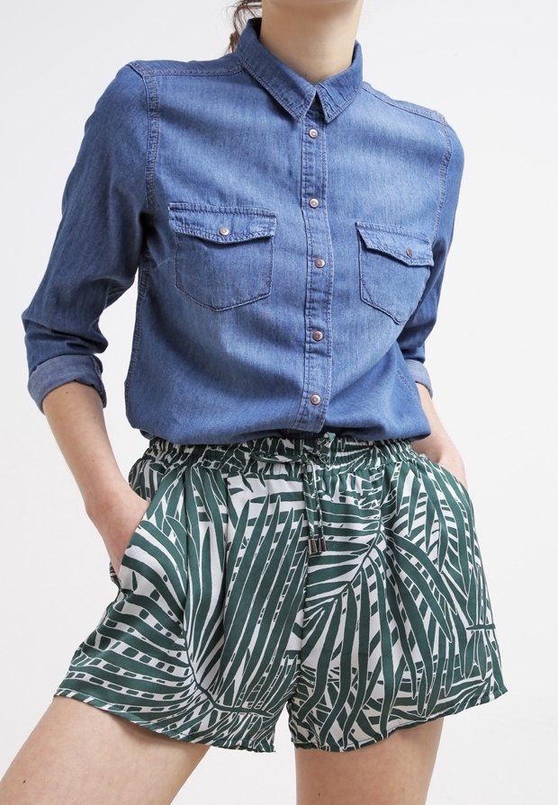 Personne portant une chemise en jean bleue boutonnée avec les manches retroussées et un short vert et blanc à motifs avec une taille à cordon, les mains dans les poches.
