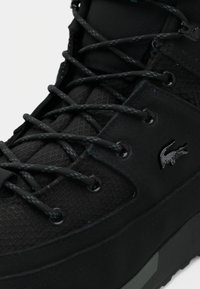 Gros plan d'une basket noire avec des lacets à motifs et un petit logo crocodile sur la face latérale.