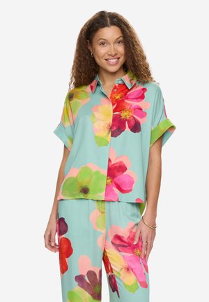 Kobieta ubrana w luźną koszulę i spodnie z dużymi, kolorowymi nadrukami kwiatowymi na jasnoturkusowym tle, uśmiechająca się do kamery.