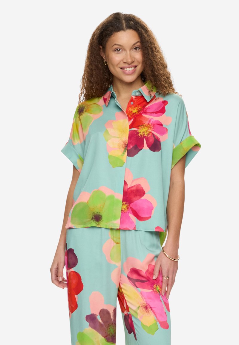 Femme portant une chemise ample et un pantalon assortis avec de grandes impressions florales colorées sur un fond turquoise clair, souriante à la caméra.