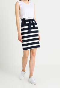 Námořnická a bílá pruhovaná sukně s páskem na zavázání, spárovaná s bílým topem bez rukávů a bílými teniskami. Hladký materiál a přiléhavý střih.