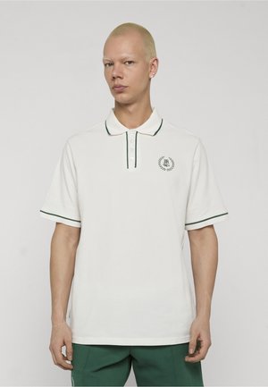 Homme aux cheveux blonds courts portant un polo blanc avec un liseré vert et un pantalon vert, debout devant un fond uni.