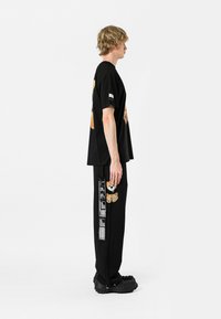 Camiseta de algodón negra con diseño gráfico de oso; combinada con pantalones negros que tienen detalles en los bolsillos y texto blanco. Zapatos negros completan el look.