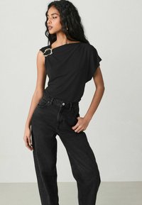 Sort, draperet kortærmet bluse med en guldfarvet nål ved skulderen, parret med høj talje sorte jeans i denim-stof.