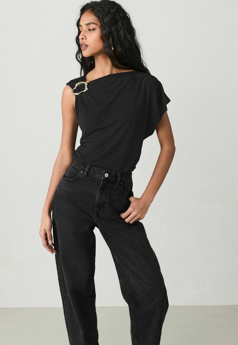 Sort, draperet kortærmet bluse med en guldfarvet nål ved skulderen, parret med høj talje sorte jeans i denim-stof.