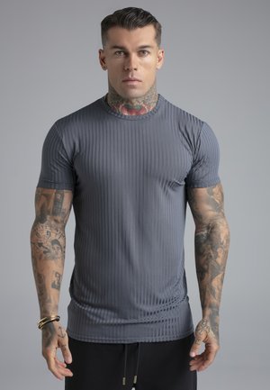 Mężczyzna z tatuażami na ramionach i szyi nosi dopasowaną, prążkowaną szarą koszulkę i czarne spodnie na jednolitym szarym tle, stoi frontem.