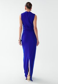 Dolga modra obleka iz gladke tkanine, s prilegajočo silhueto, naborki na hrbtu in visokim razporkom na stegnu. Vidni so zlati sandali z rametnicami.