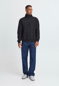 Sweat à zip noir avec un col haut, associé à un jean en denim bleu et des baskets grises. Design simple, coupe décontractée, texture douce.