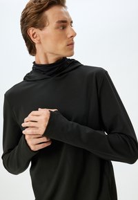 Top negro de manga larga con capucha, confeccionado con una tela suave y elástica. Los orificios para los pulgares en los puños mejoran la funcionalidad y la comodidad. Diseño minimalista.