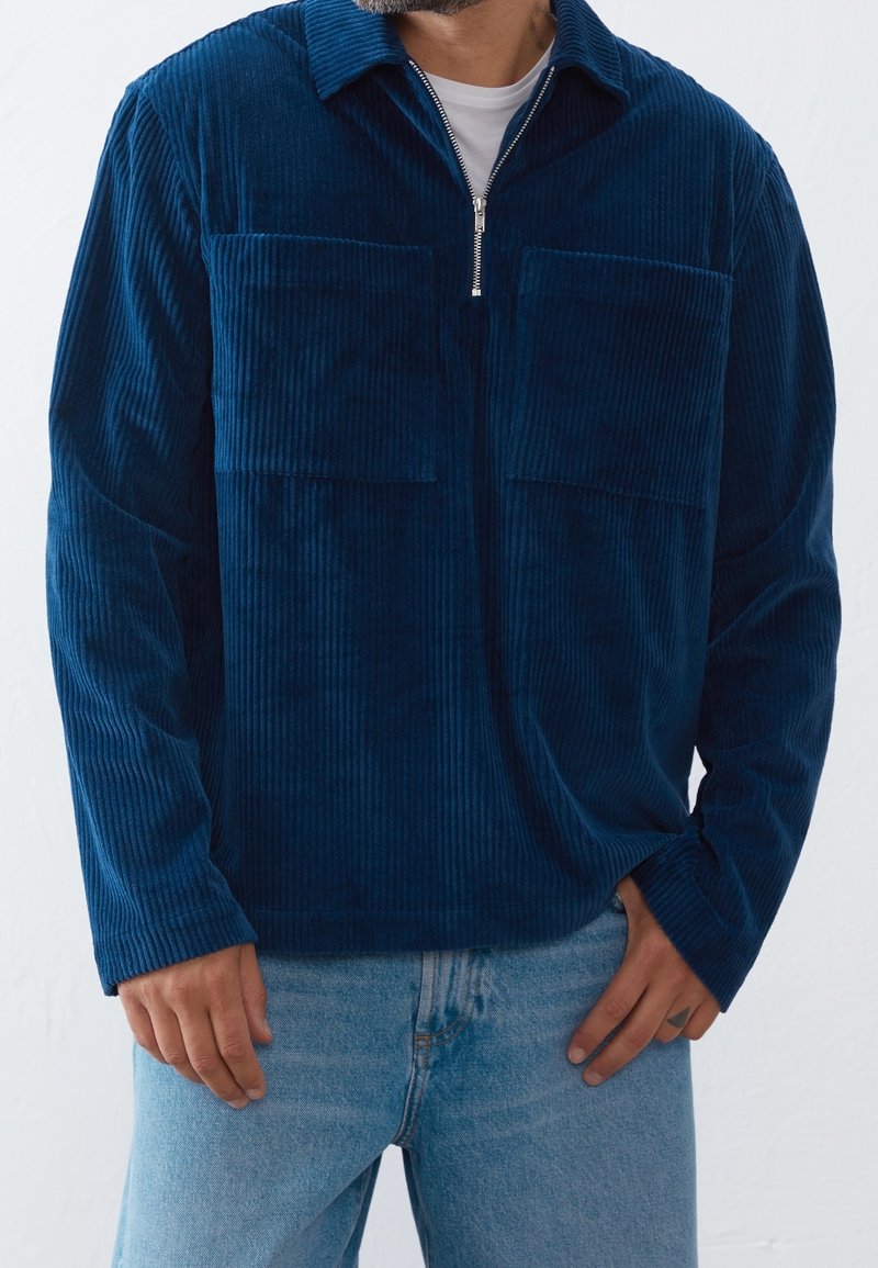Homme portant un pull à fermeture éclair en velours côtelé bleu avec de grandes poches poitrine et un jean bleu clair, la main reposant près de la poche.
