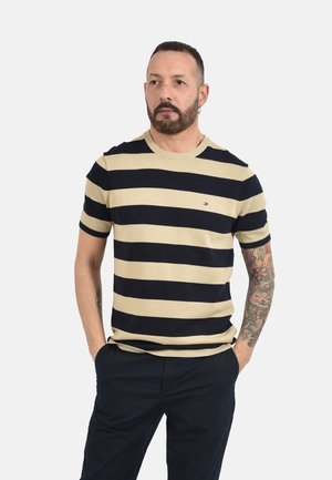 Uomo con barba e braccio tatuato che indossa una maglietta beige e nera a righe a maniche corte e pantaloni scuri, in piedi con le mani nelle tasche.