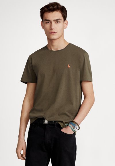 Olive green cotton t-shirt with a crew neck, featuring a small embroidered logo on the left chest, paired with black jeans and colorful bracelets.