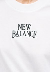 Λευκό βαμβακερό t-shirt με μαύρα κεντημένα γράμματα που αναγράφουν «NEW BALANCE» με κυρτή, έντονη γραμμ ατοσειρά. Λείο υφή και στρογγυλή λαιμόκοψη.