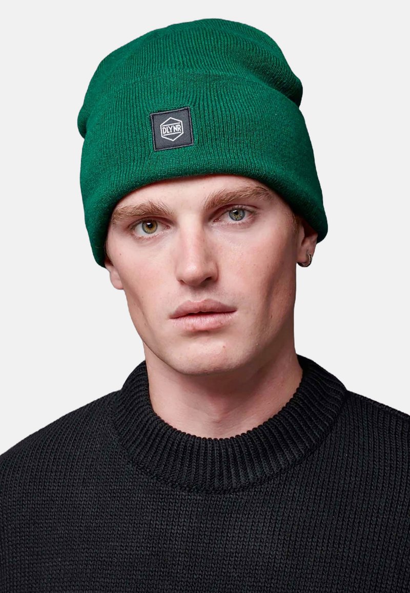 Groene gebreide beanie met ribbeltextuur en een zwart logo-patch. Draagt een zwarte ribbeltrui met een hoge hals.