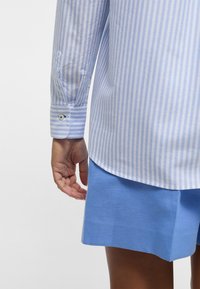 Hemd in hellblau-weißer Streifenoptik mit Knopfmanschette, kombiniert mit einer einfarbigen blauen Shorts. Das Hemd hat einen abgerundeten Saum und besteht aus weichem Stoff.