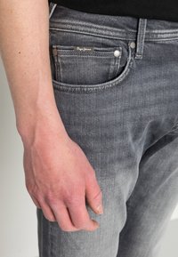 Detail ruky spočívající vedle šedých džínů z denimu s viditelnou značkou Pepe Jeans na malém předním kapse.