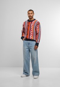 Bunter gestrickter Pullover mit verschiedenen Mustern in Rot, Blau und Gelb. Kombiniert mit hellblauen weit geschnittenen Jeans und weißen Turnschuhen.