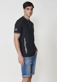 Melns polo krekls ar teksturētu apdari, pogu apkakli un baltu grafisko druku sānos. Valkāts kopā ar gaiši ziliem džinsu šortiem.