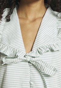 Haut rayé vert et blanc avec un lien à l'avant, un col volanté et des détails en boutons. Le tissu semble doux et léger.