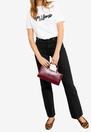 Femme portant un t-shirt blanc "Milano" et un jean noir, tenant un sac à main bordeaux brillant, et portant des ballerines imprimé léopard.