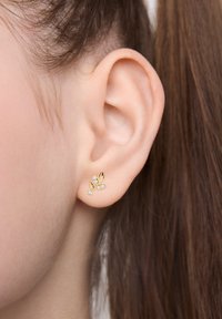 Złote kolczyki w kształcie liści z trzema małymi przezroczystymi kamieniami, o gładkiej teksturze. Noszone na małżowinie uszu modelki, z włosami związanymi do tyłu.