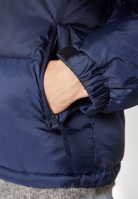 Hånd i lynlåslomme på en mørkeblå dunjakke med justerbar velcrolukning på ærmet.