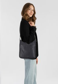 Donna con capelli mossi che indossa una giacca nera e jeans chiari, porta a tracolla una borsa in pelle blu scuro, sorridendo leggermente, su sfondo bianco.