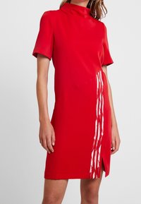 Röd klänning med hög krage och korta ärmar. Har en asymmetrisk design med ett vitt vertikalt rand och slitsar i fållen.
