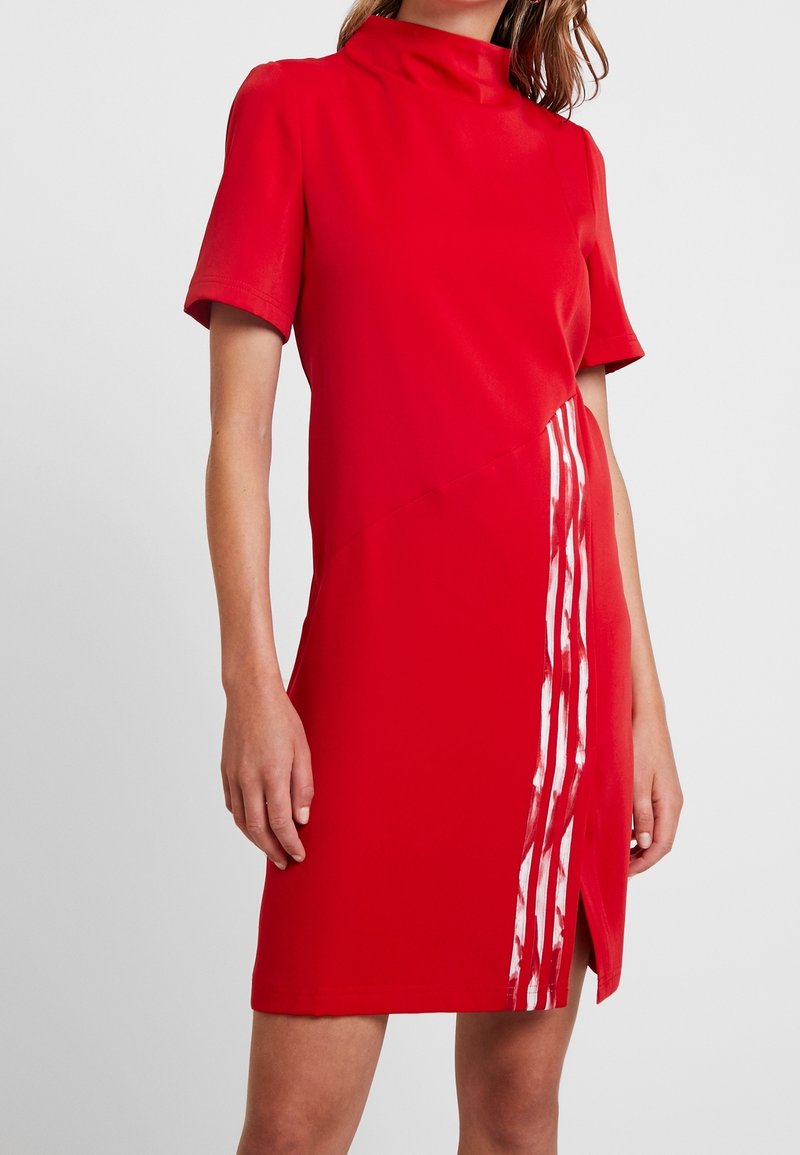 Röd klänning med hög krage och korta ärmar. Har en asymmetrisk design med ett vitt vertikalt rand och slitsar i fållen.