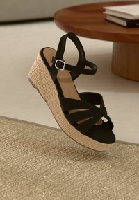 Sandale noire à talon compensé avec plusieurs sangles et une boucle en argent. Caractérisée par un talon en jute tissé. Semelle lisse avec un fond texturé.
