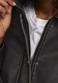 Giubbotto di pelle nera con zip visibile e fodera bianca, caratterizzato da una superficie testurizzata e dettagli con bottoni a pressione. T-shirt bianca sotto.