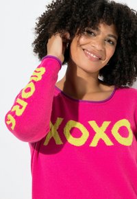 Lächelnde Frau mit lockigem Haar, die einen leuchtend rosa Pullover mit gelber "XOXO"-Schriftzug auf der Brust und am Ärmel trägt.