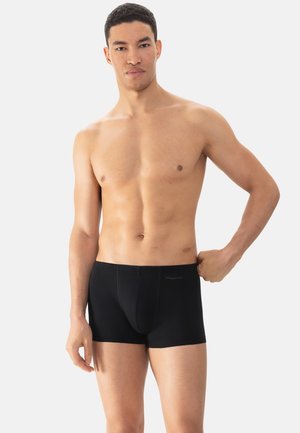 Mężczyzna bez koszulki w czarnych slipach stoi na jednolitym tle, jedną ręką szarpiąc za pasek po prawej stronie.