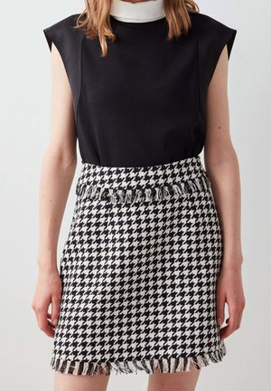 Woman wearing sleeveless black top with white collar and black-and-white houndstooth skirt with fringed edges, standing against plain background.