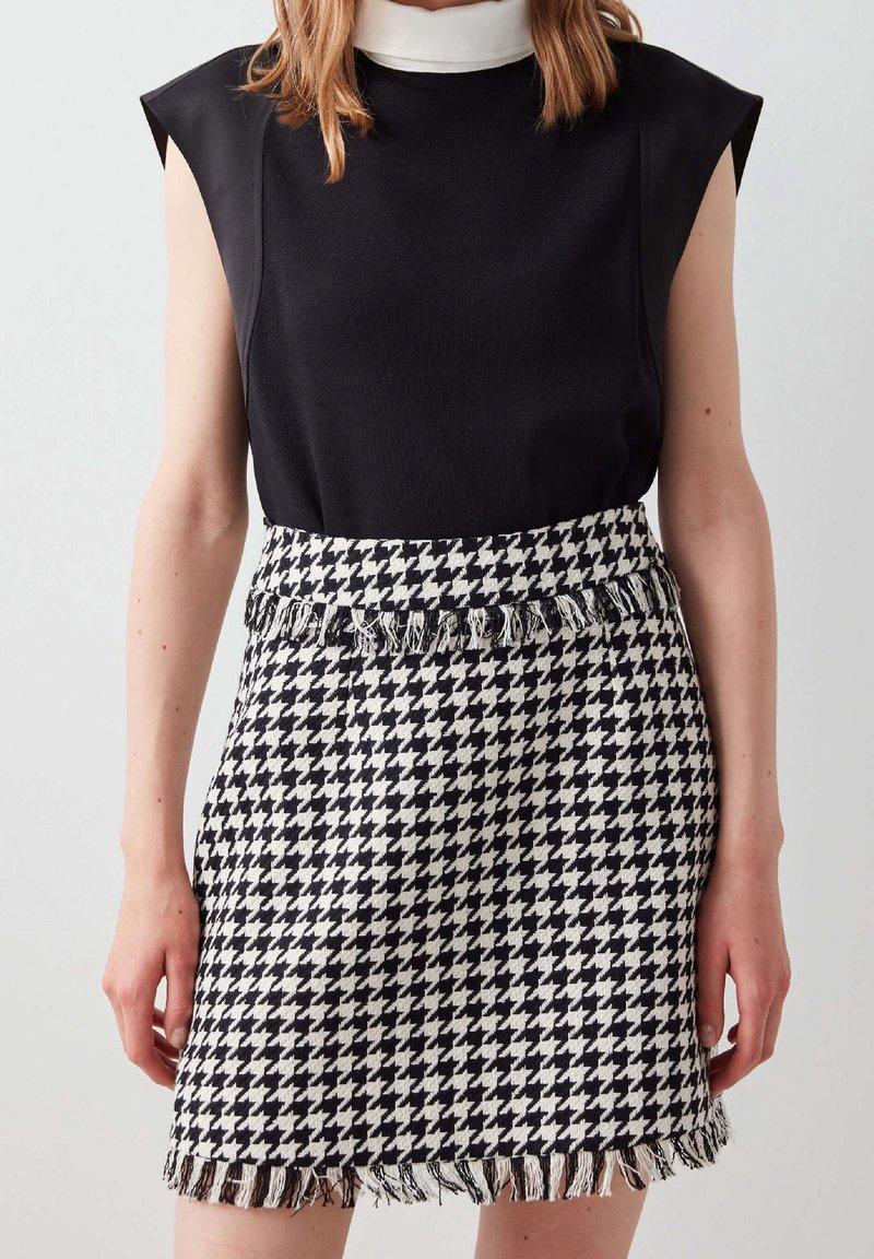 Femme portant un haut noir sans manches avec un col blanc et une jupe pied-de-poule noire et blanche à bords frangés, debout devant un fond uni.