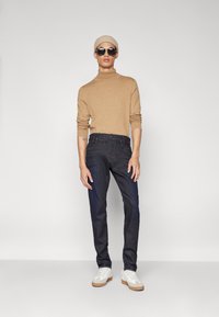 Suéter de cuello alto beige, jeans oscuros y zapatillas blancas. La modelo lleva gafas de sol y un gorro de punto beige. Silueta simple y moderna.
