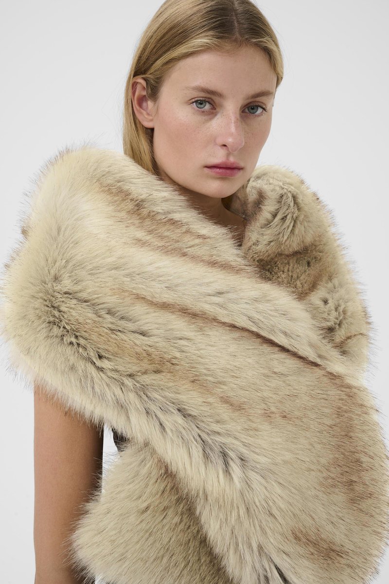 Giovane donna con capelli biondi che indossa uno scialle in pelliccia sintetica a strisce beige e marroni su uno sfondo bianco.