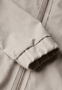 Beige jakkeærme med en sammensnerpet manchet, tekstureret stof og en synlig metallynlås langs sømmen. Blødt, glat udseende.