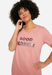 Camiseta de color rosa claro con mangas cortas, que presenta letras metálicas que dicen "BUENA KARMA". Material suave y corte relajado.