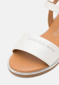 Baltos plokščios šlepetės su sintetiniu dirželiu, ant kurio iškilmingai įspaustas "Calvin Klein Jeans" logotipas, tan spalvos paminkštinta padas ir sidabriniai akcentai palei kraštą.