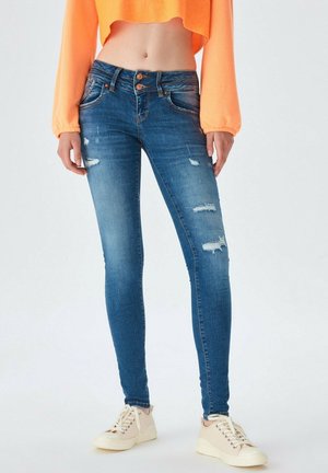 Femme portant un jean skinny bleu déchiré avec une fermeture à double bouton, un haut orange à manches longues raccourci, et des baskets beiges, debout.