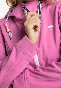 Rosa hoodie med hög krage, dragsnoddar och en liten broderad paraplylogotyp. Tillverkad av ett mjukt material med en slät textur.