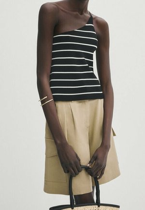 Personne portant un haut asymétrique noir et blanc à rayures, un short beige, des bracelets en or, tenant un sac tissé à poignées noires.