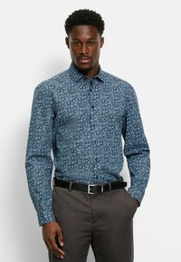 Button-up-Hemd aus blauem Stoff mit floralem Muster. Lange Ärmel mit Knopfmanschetten. Kombiniert mit dunklen Hosen und einem schwarzen Gürtel.